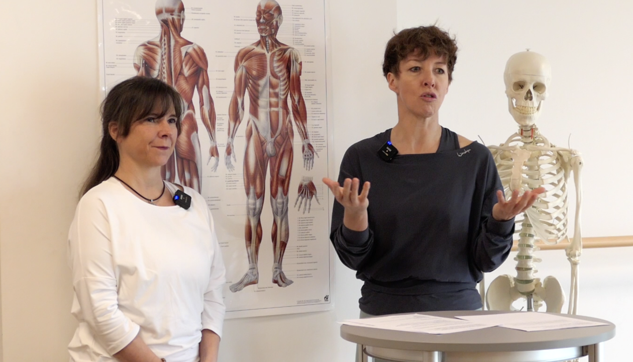 Jenni Bergunde und Kerstin Bredehorn stehen an einem Stehpult, hinter ihnen is ein Plakat mit einer anatomischen Zeichnung der Muskeln am Menschen.