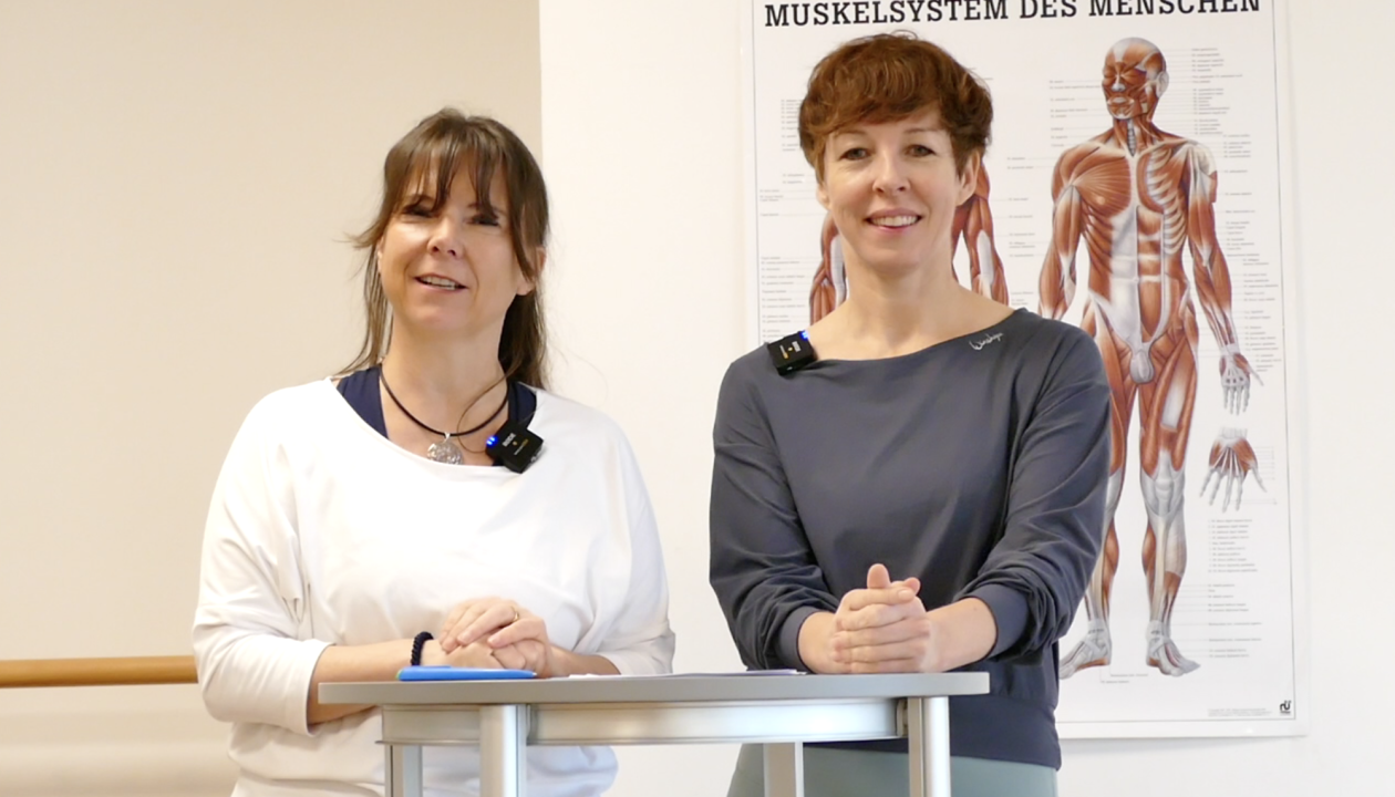 Kerstin Bredehorn und Jenni Bergunde stehen an einem Stehtisch, hinter ihnen hängt ein Plakat mit dem Muskelsystem des Menschen.