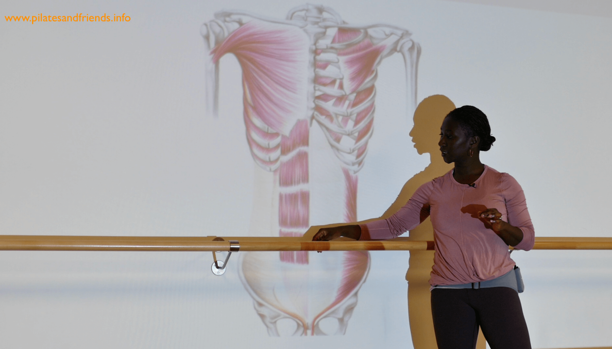 Juliana Afram steht vor einer Ballettstange, hinter ihr ein projeziertes Bild von Brustkorb und Bauchmuskeln