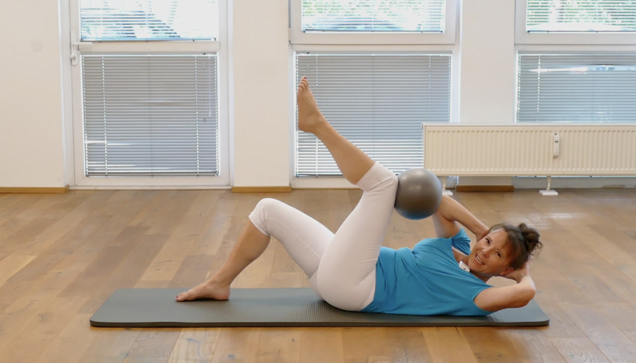 Kerstin Bredehorn in Rückenlage auf einer Matte, führt die Übung crisscross aus mit dem Ball zwischen Ellenbogen und Knie